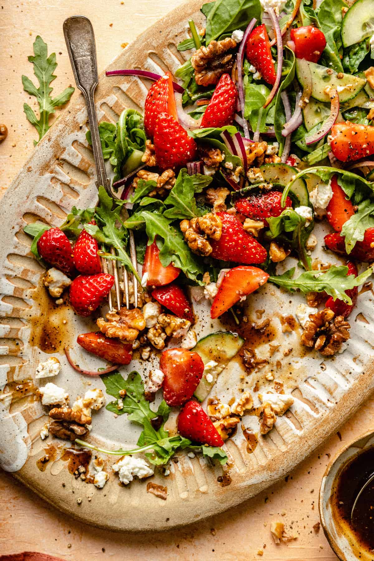 A messy platter showing some of the strawberry salad having been served up, a fork and bowl of dressing to the side.