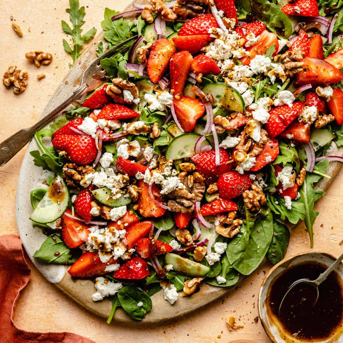 A platter with strawberry walnut salad served on it with a fork and bowl of balsamic dressing.