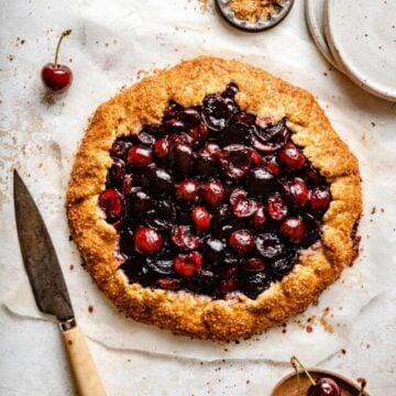 Cherry galette on parchment with a knife and cherries around with plates.