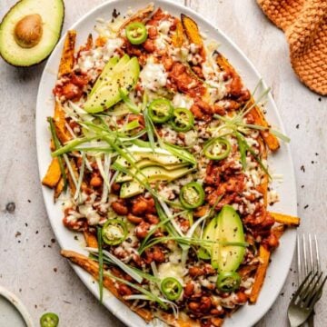 Loaded sweet potato fries on a platter with avocado, a fork and napkin nearby.