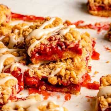 A stack of strawberry crumble bars with crumbs around and a creamy glaze drizzled on top.