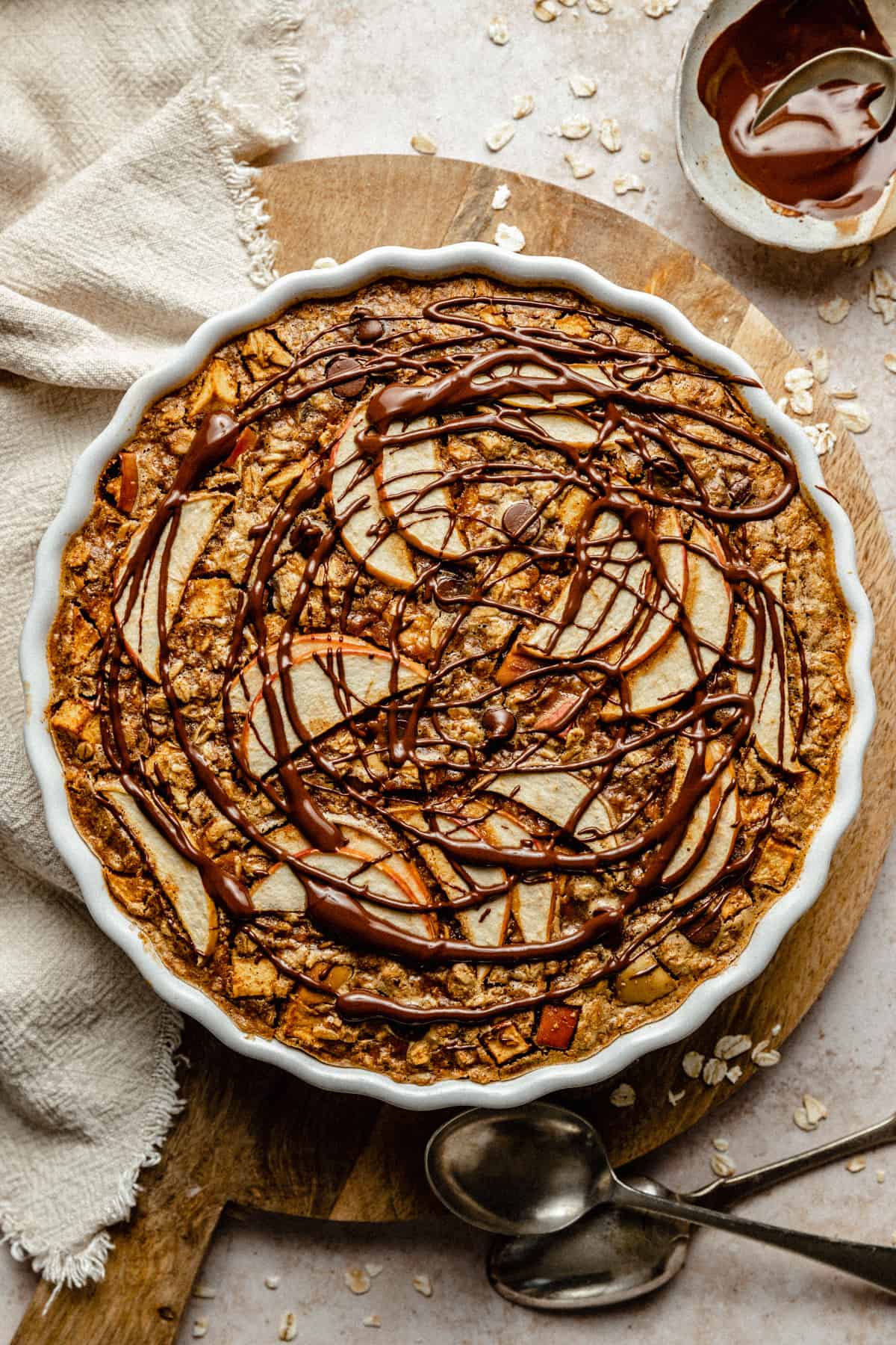 An apple topped baked oatmeal in a white baking dish on a wooden board with a napkin nearby.