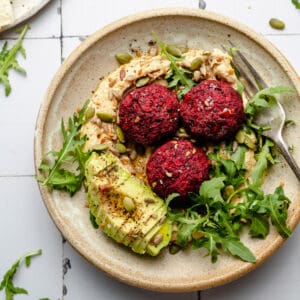 Beetroot falafel in a bowl served on top of hummus with avocado and leaves and a fork.