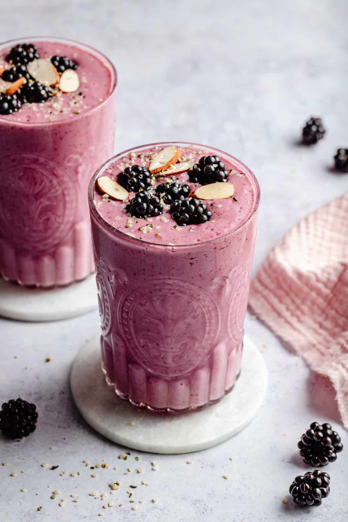 Two glasses of blackberry strawberry banana smoothie on marble coasters with fruit and seeds on top.