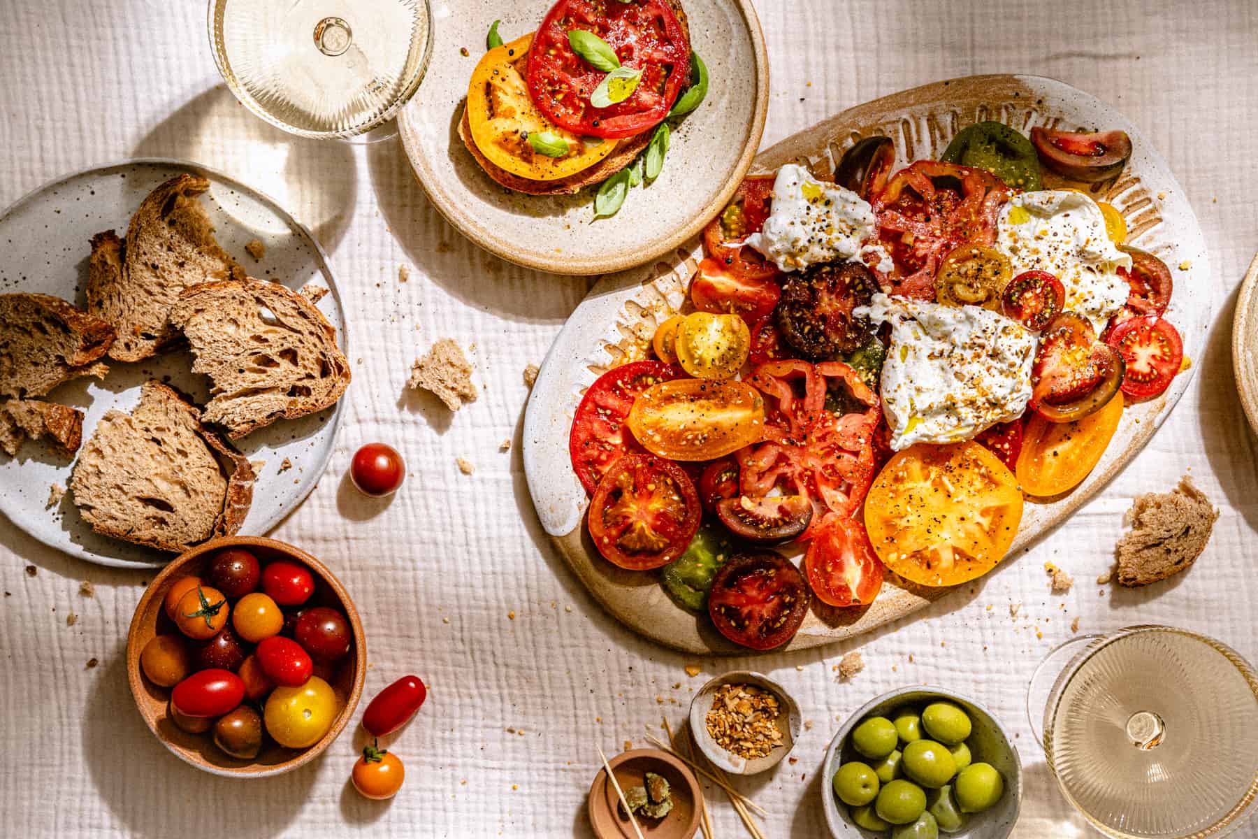 A table with platters of tomatoes, bread, burrata and olives with glasses of wine.
