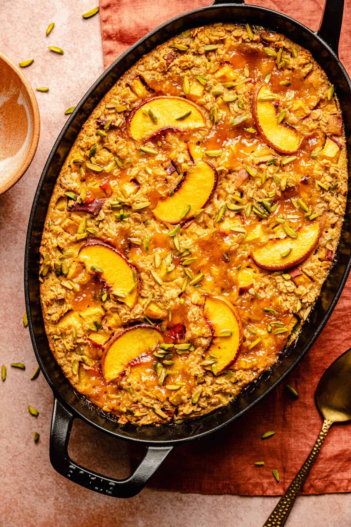 A peach baked oatmeal in a black casserole dish with a golden spoon to the side.