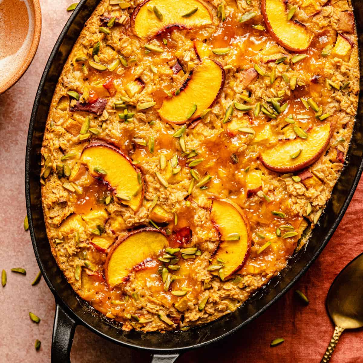 A close up of a cooked peach baked oatmeal in a baking dish with a bowl and spoon to the side.