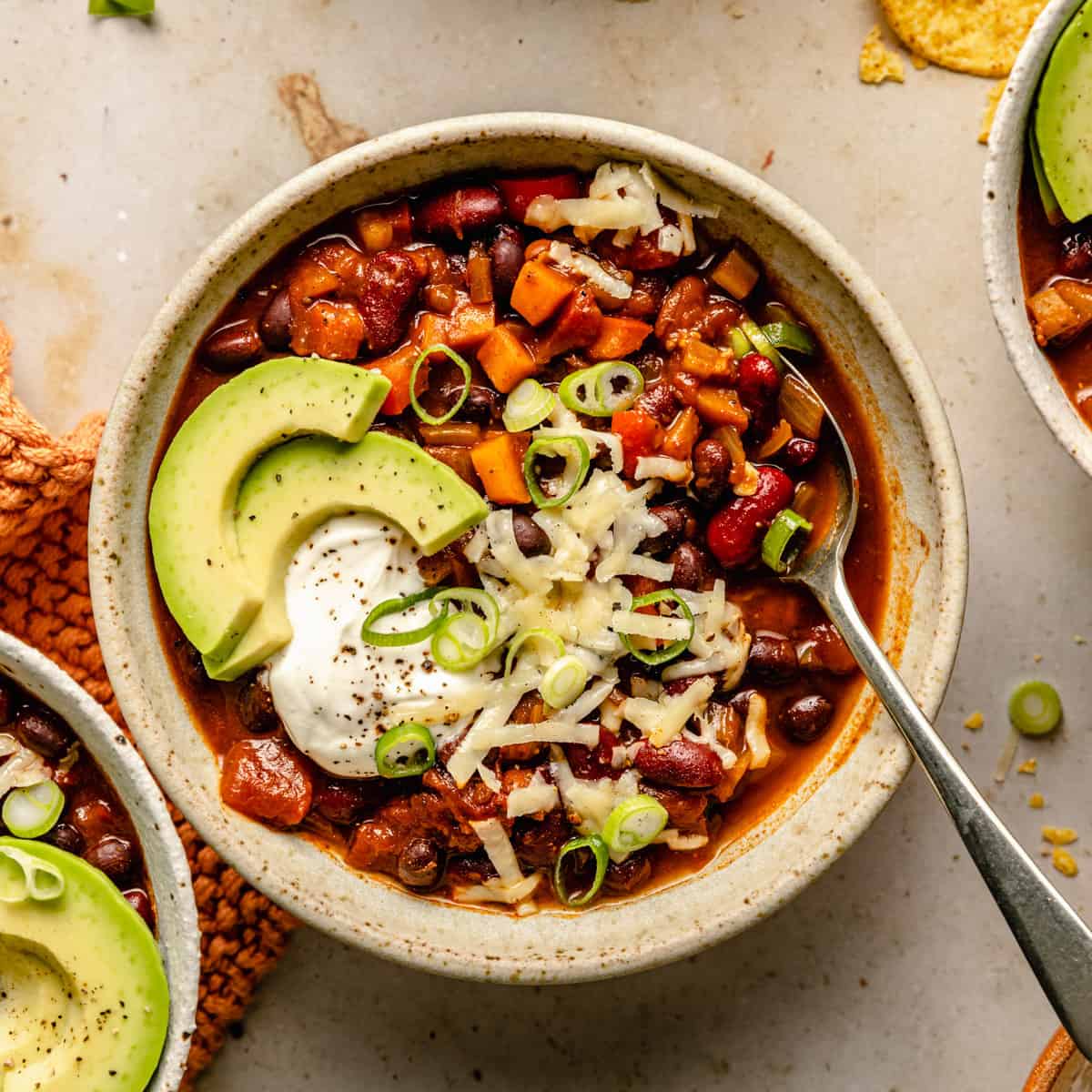A bowl of three bean chili with cheese, sour cream and avocado on top with a spoon.