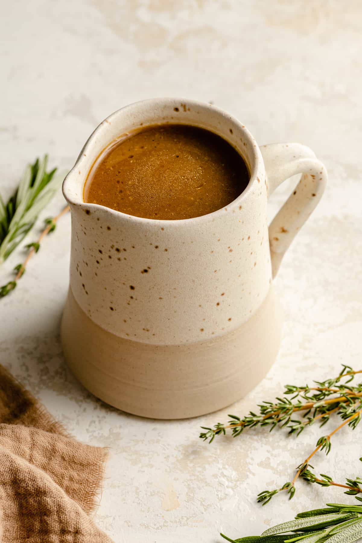 A jug of chicken gravy with a napkin and herbs around by.