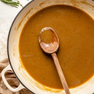 Chicken gravy in a skillet with a spoon and herbs around.