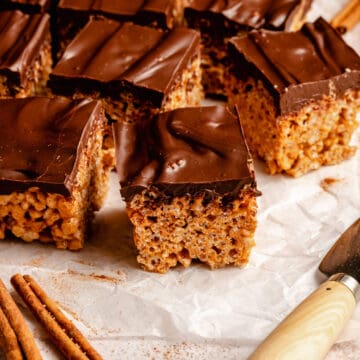Chocolate covered rice krispie treats cut into squares on parchment with a knife and cinnamon sticks.