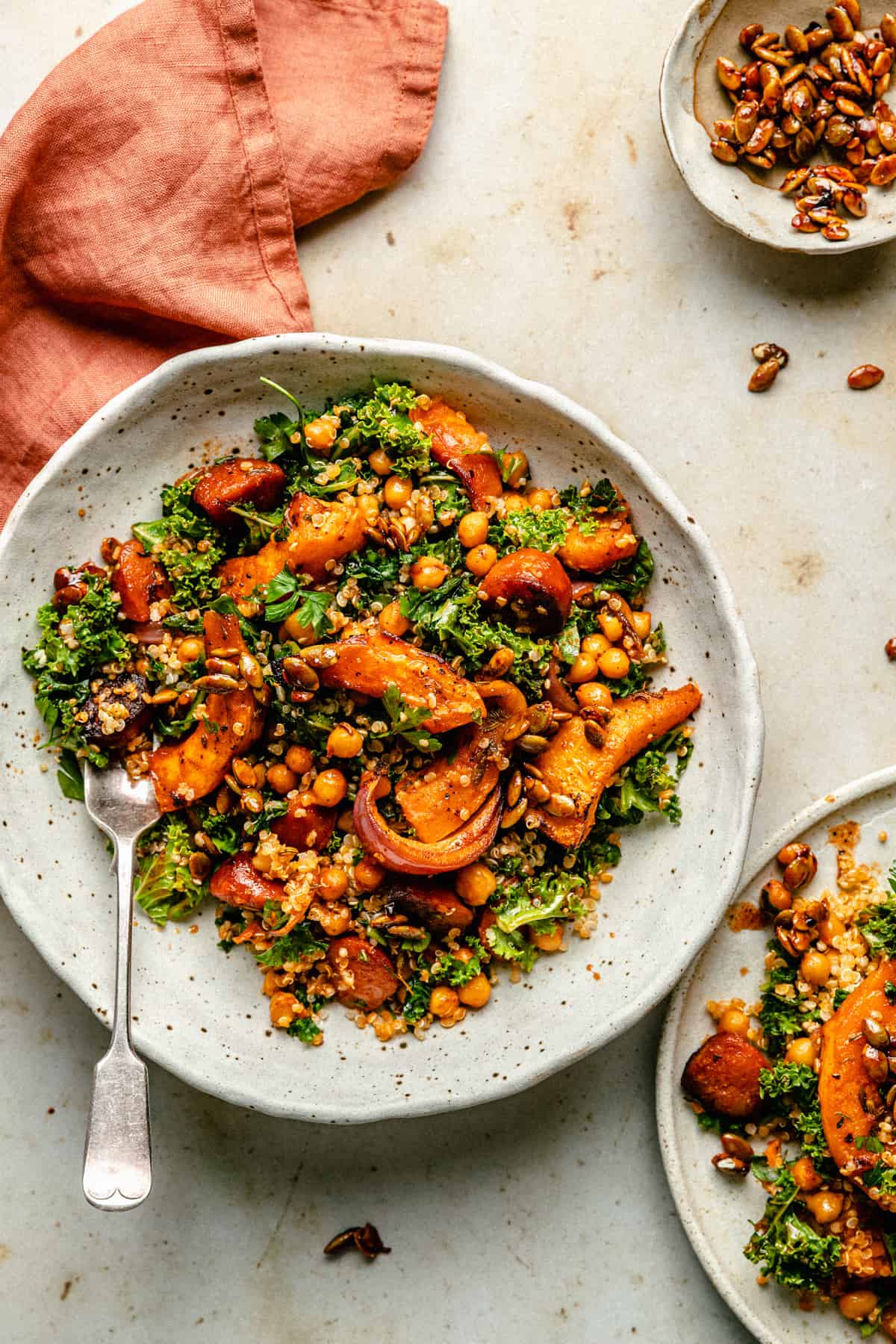 A cream bowl of roast pumpkin salad with a fork, toppings and a napkin to the side.