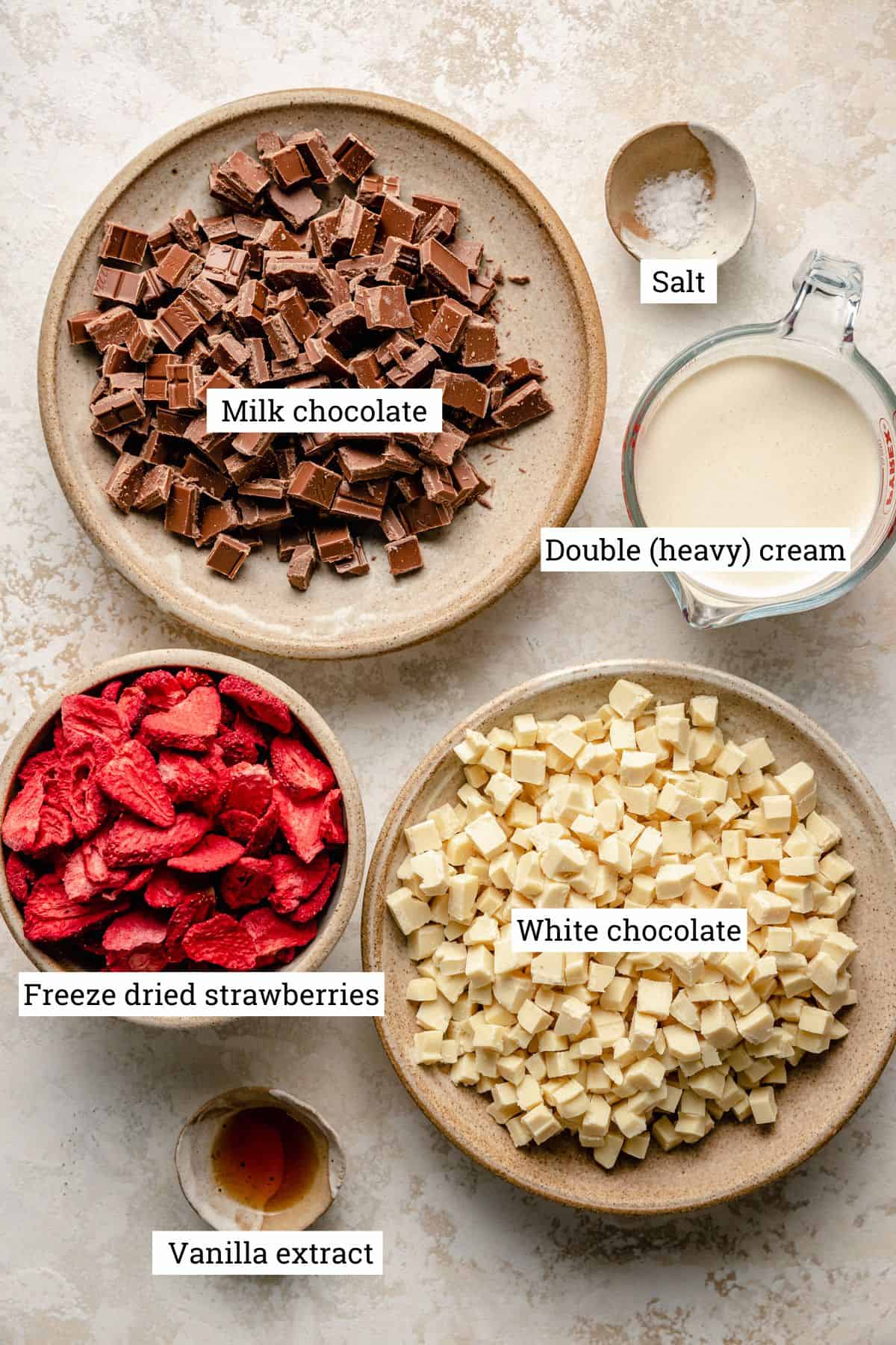 Ingredients in bowls on a work surface including chocolate, heavy cream and freeze dried strawberries.