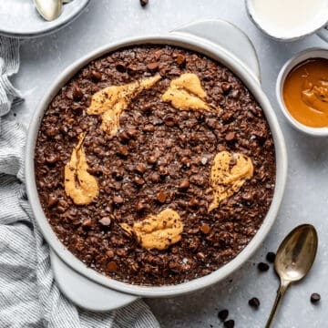 Chocolate peanut butter baked oatmeal in a dish surrounded with bowls, peanut butter, spoon and milk.