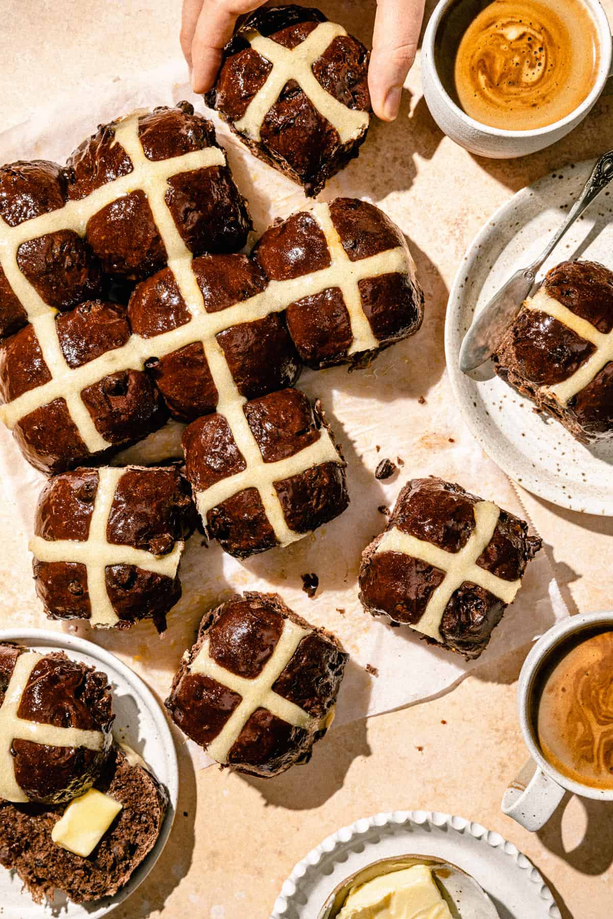 An array of hot cross buns on parchment paper with a coffee and plate to the side.