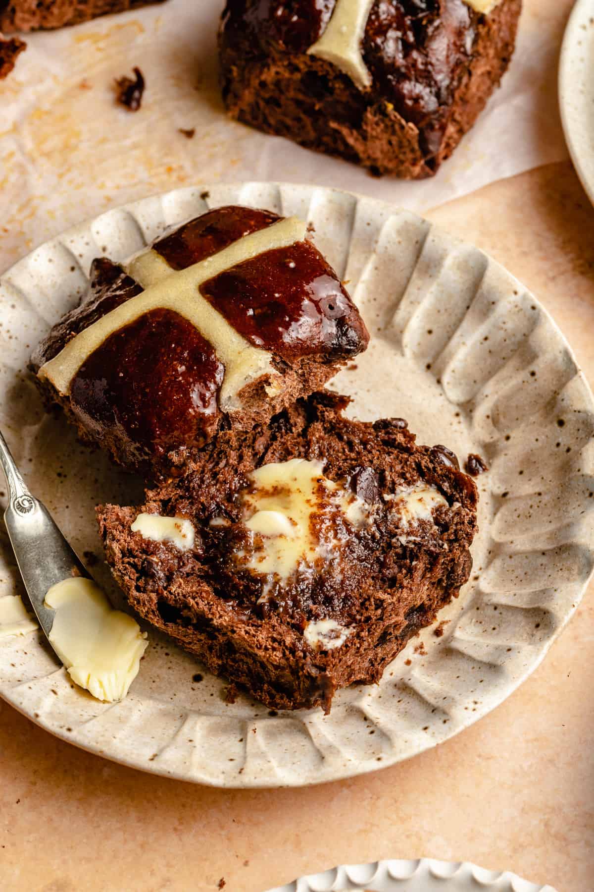 A cut open chocolate hot cross bun on a plate with a knife and butter spread on it.