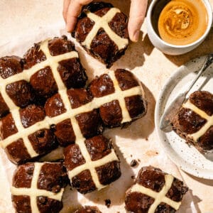 Chocolate hot cross buns in an array with a hand picking up on and a coffee to the side.