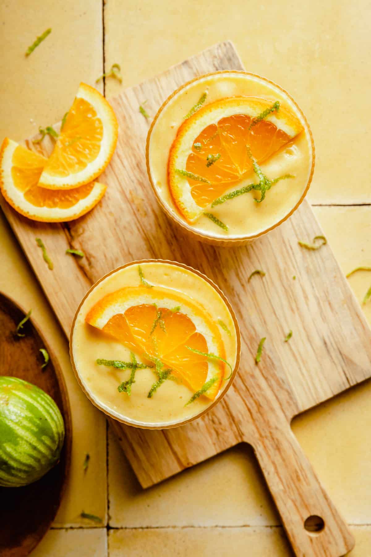 Overhead of two glasses of orange mango smoothie on a wooden board with orange slices around.