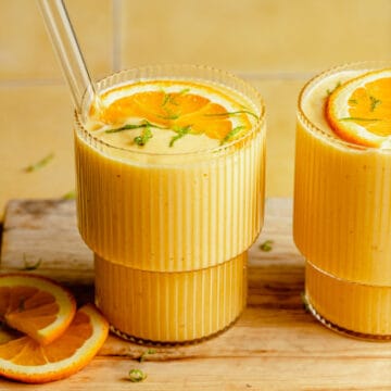 Two glasses of orange mango smoothie with a glass straw on a wooden board.
