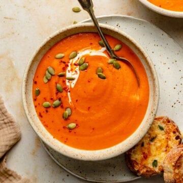 A bowl of butternut squash and red pepper soup with yogurt and pumpkin seeds on top with a spoon.