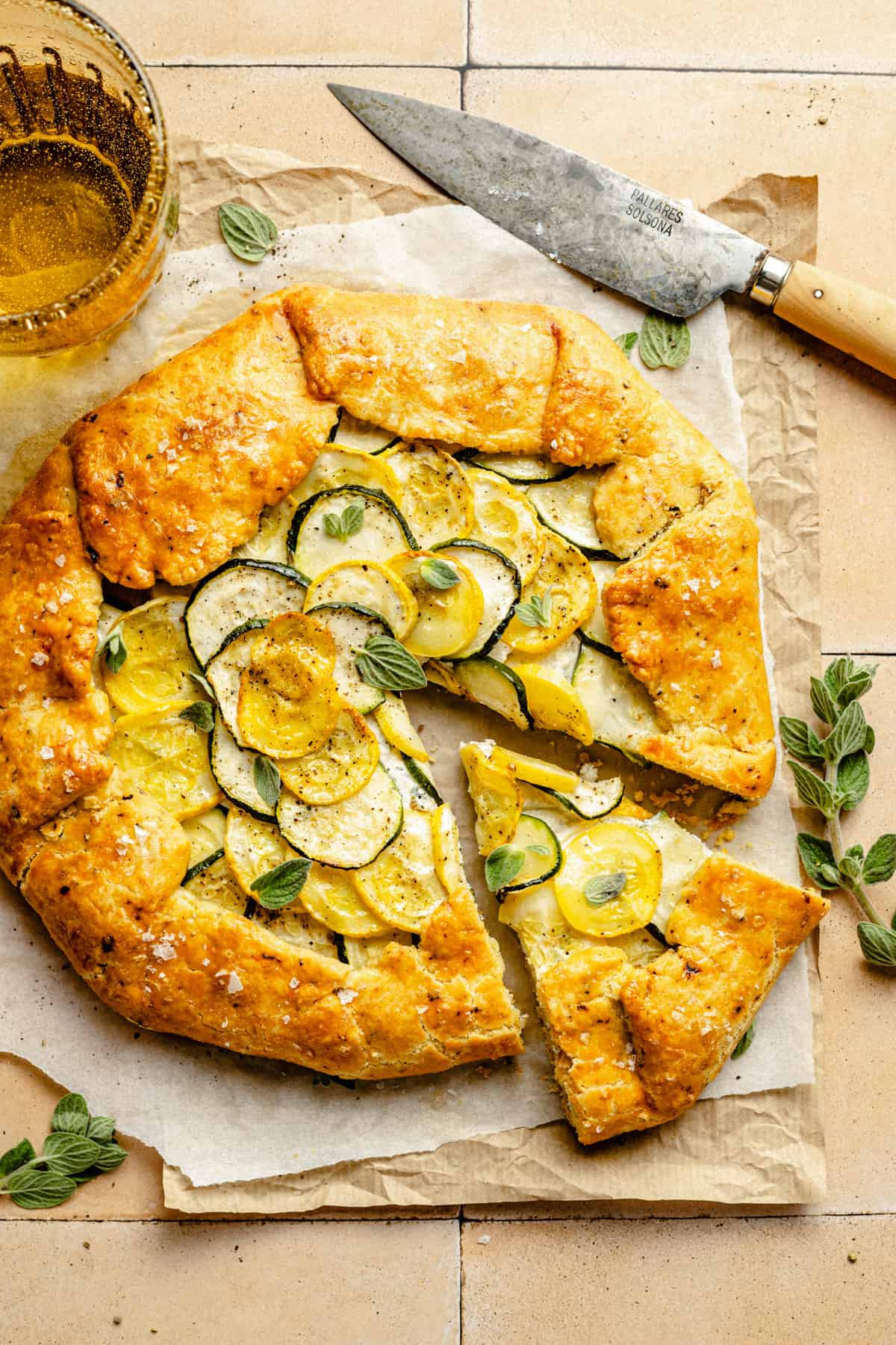 The zucchini galette with a wedge cut, knife to the side with herbs and a drink too.