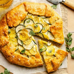 Zucchini galette on parchment with a wedge cut out showing a golden crust and a knife to the side.