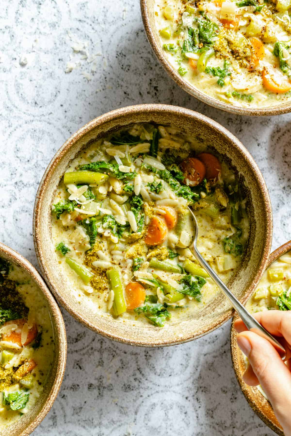 A bowl of vegetable orzo soup with a hand holding a spoon.