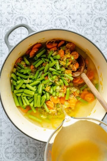 Pouring vegetable broth into the vegetable orzo mixture in a dutch oven.