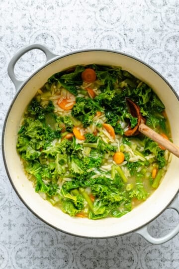 Stirring in kale to the vegetable orzo soup with a wooden spoon.