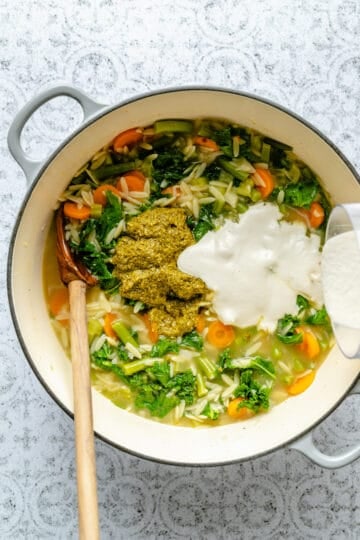 Cashew cream being poured into the the vegetable orzo soup in a dutch oven with a wooden spoon.