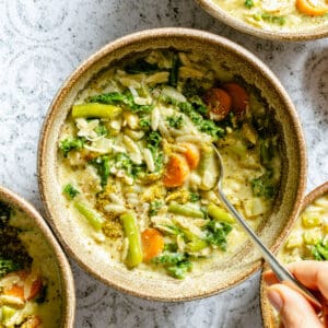 A bowl of creamy pesto vegetable orzo soup with a hand holding a spoon in the bowl.